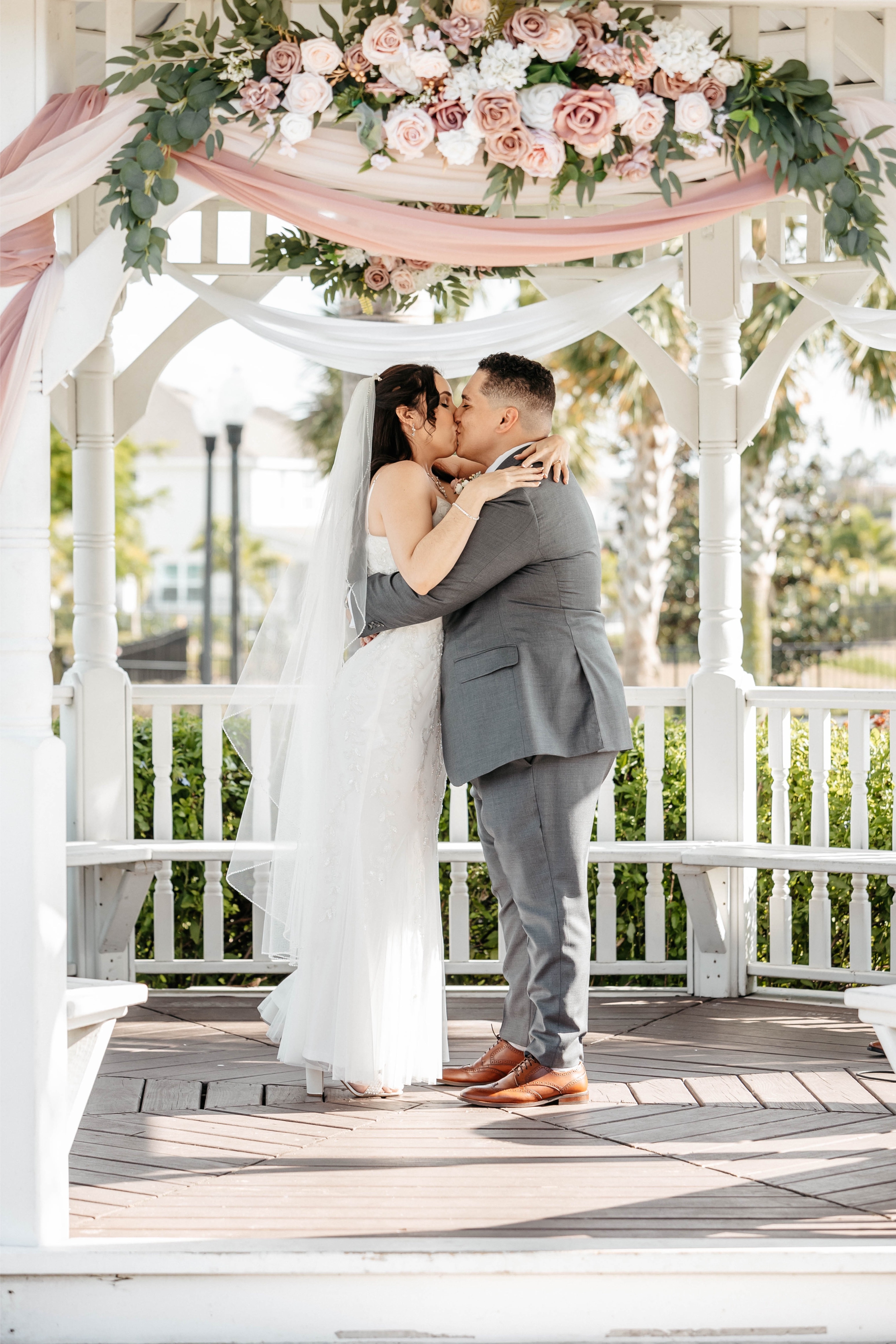 A groom kisses his bride under a gazebo decorated with white draped linen and pink and white flowers at a wedding venue in Central Florida