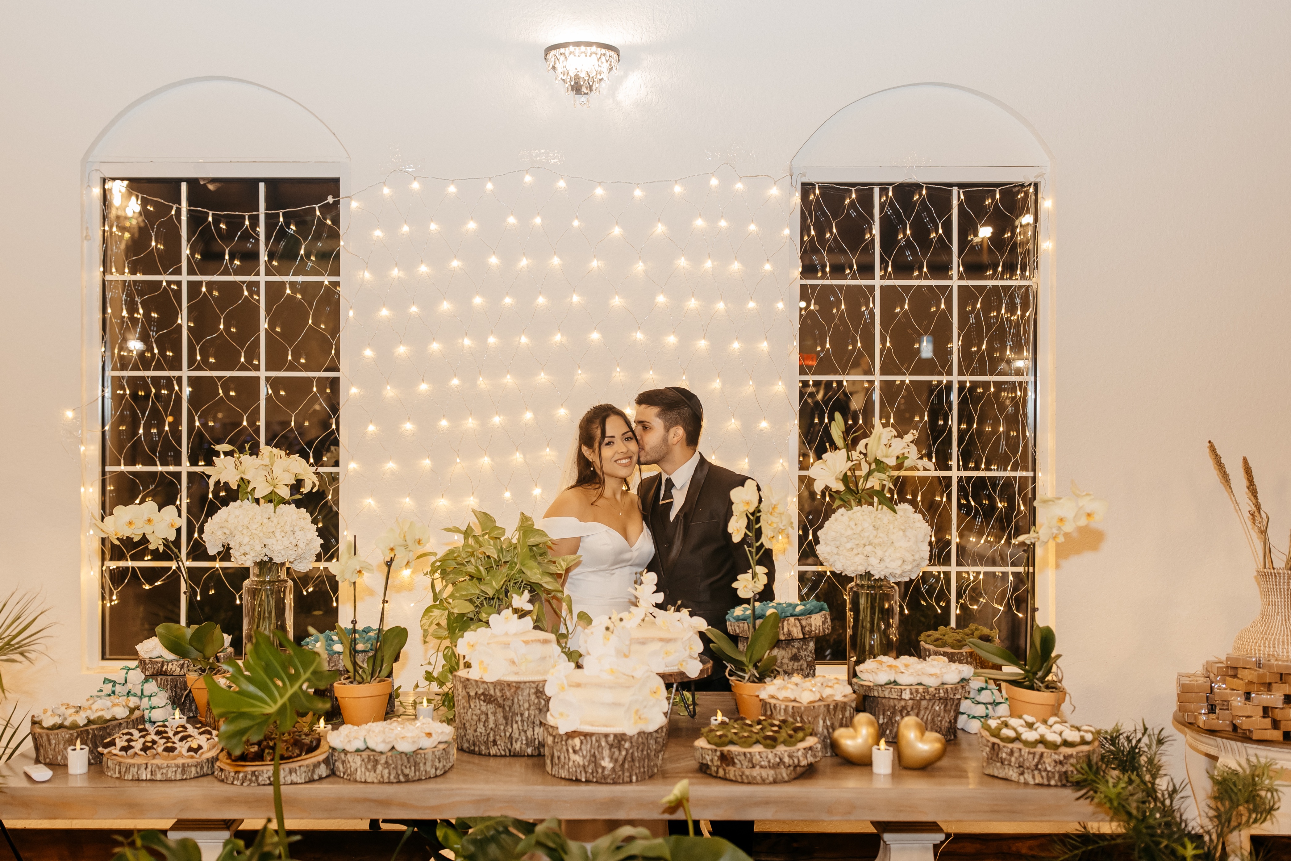 A bride and groom stand behind a gorgeous spread of treatrs and cakes with white flowers and greenery at one of the affordable wedding venues in central florida 
