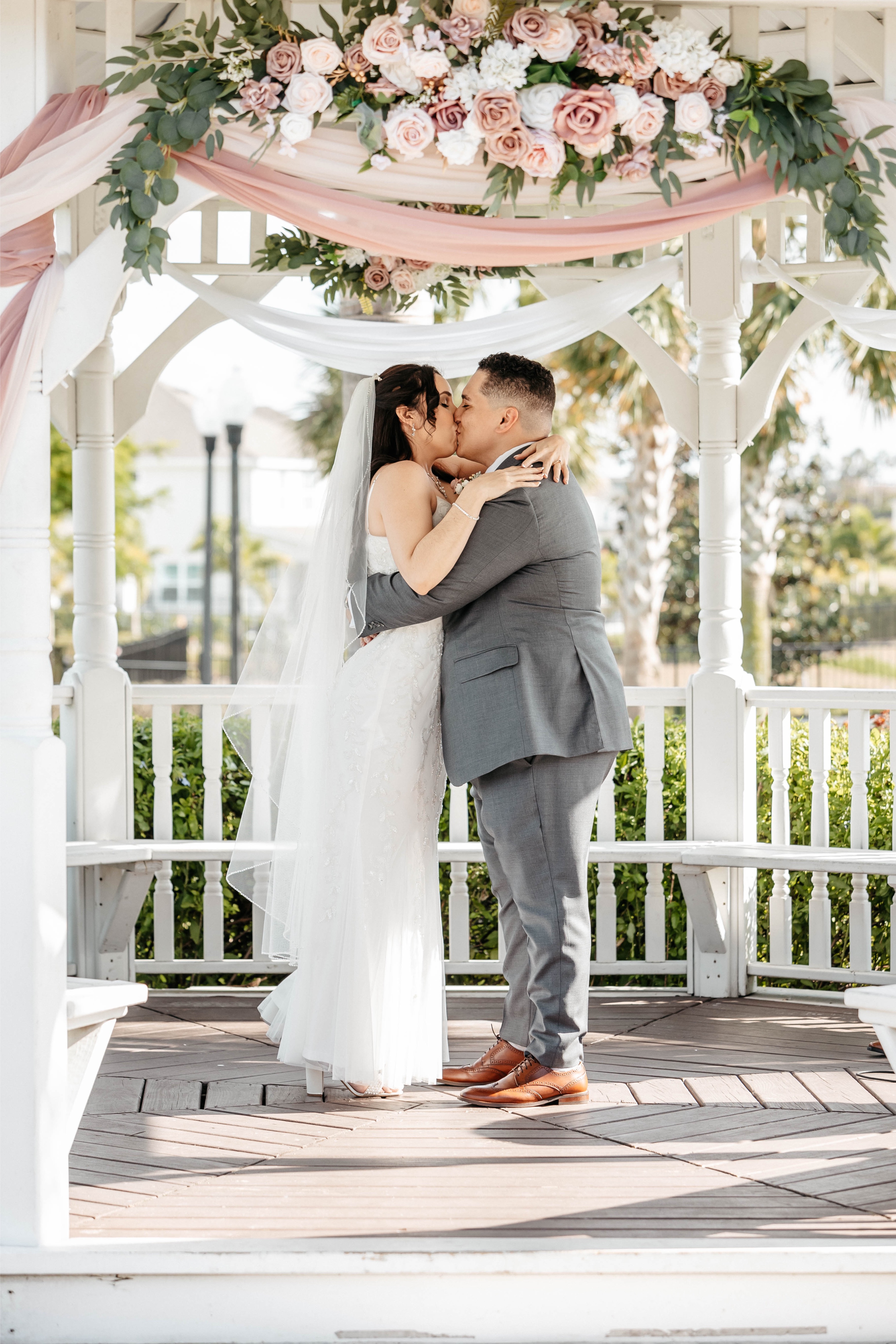 A groom kisses his bride under a gazebo decorated with white draped linen and pink and white flowers at a wedding venue in Central Florida 