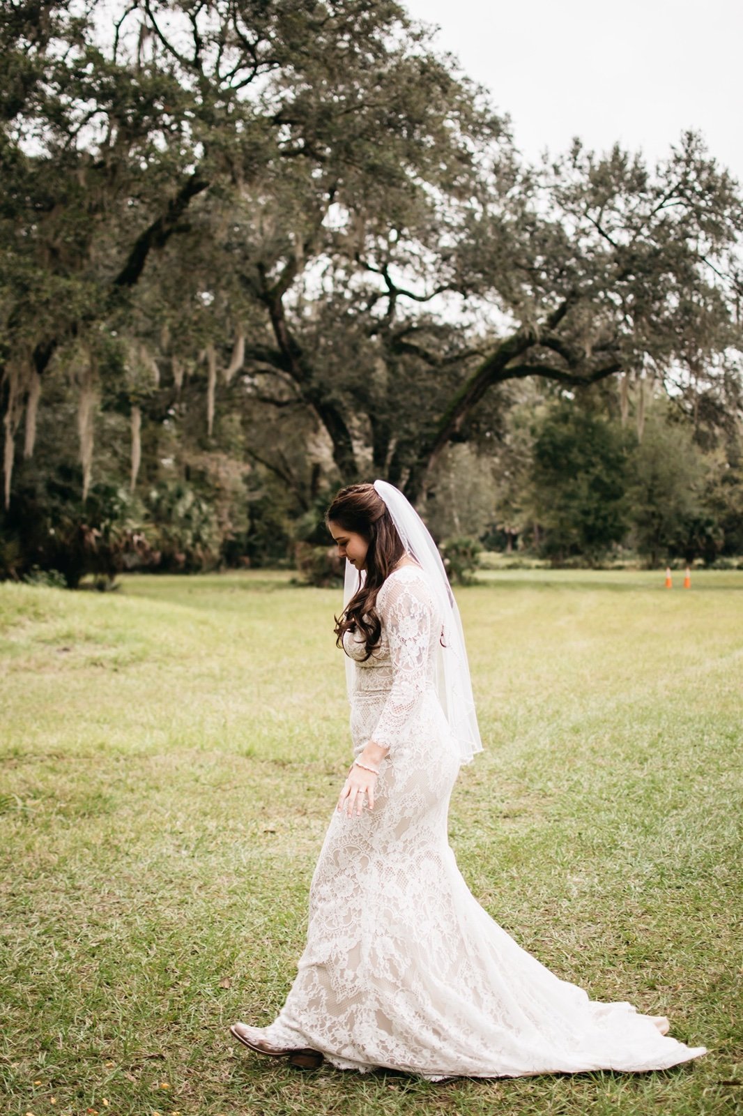 A bride in a lace wedding dress and veil walks towards her groom in a field surrounded by large trees at a ​wedding venue in bushnell fl
