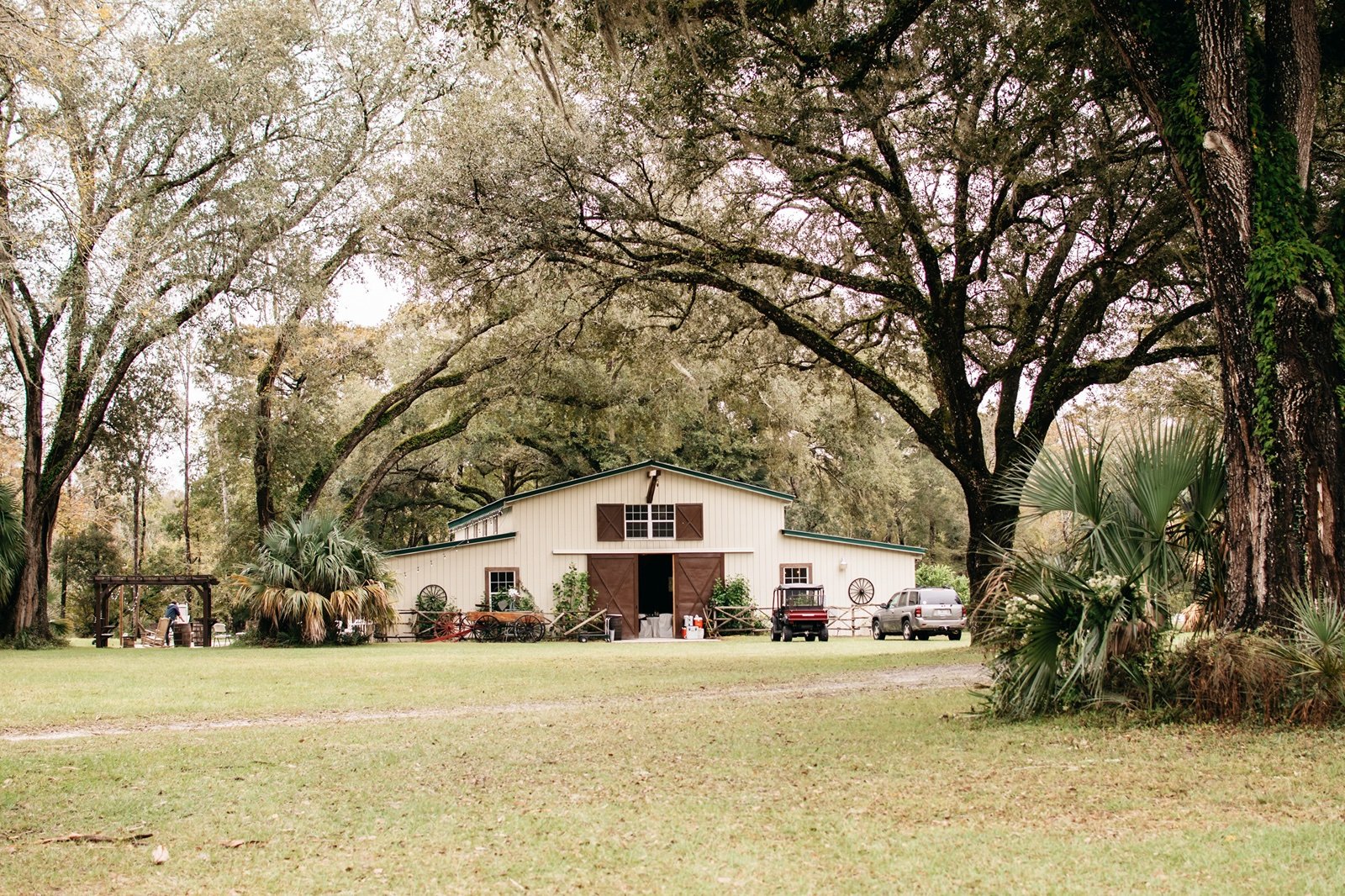 A white barn with brown doors and a green roof sits in a field surrounded by large, looming trees awaiting to be used for a 
bushnell wedding
