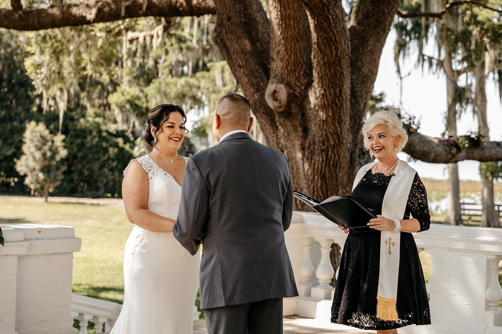 A bride and groom hold hands while their officiant smiles and speaks at their bella cosa wedding
