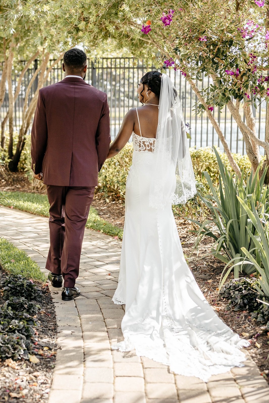 A bride and groom wearing a burgundy suit walks down a brick path through a garden in outdoor wedding venues in central florida