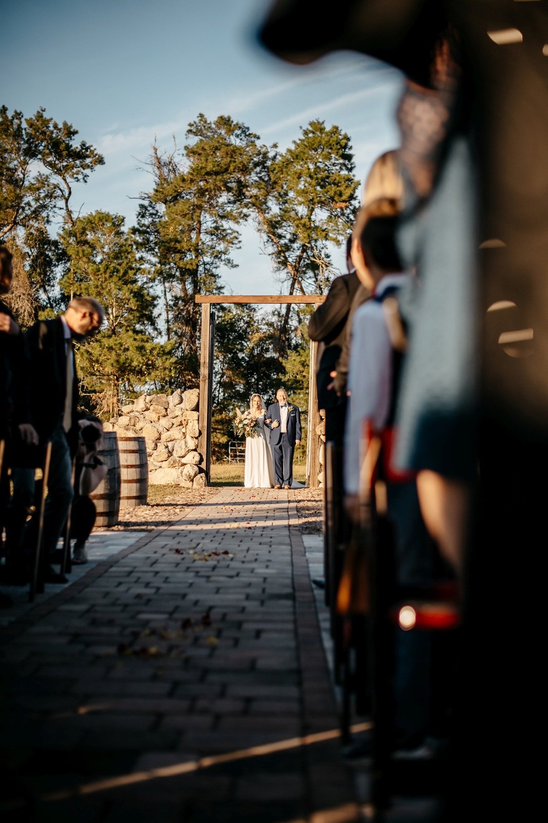 A bride and her father walk down a brick path to her groom passing by her excited guests at woodsy outdoor wedding venues central florida
