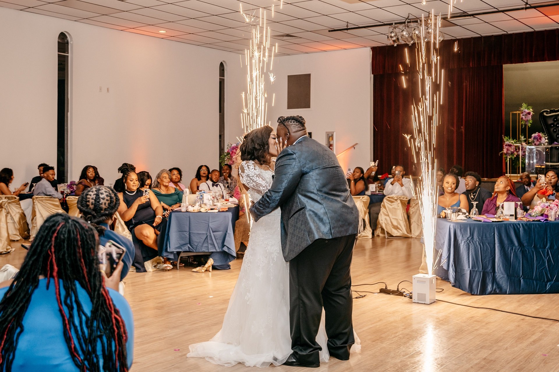 A bride and groom kiss on the dance floor at their Central Florida wedding with fireworks in the background while family and friends cheer.